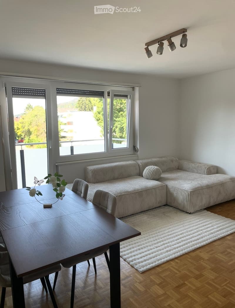 Modern living room with a sectional sofa, wooden dining table, chairs, and a white shag rug on a wooden floor. Large windows provide natural light.
