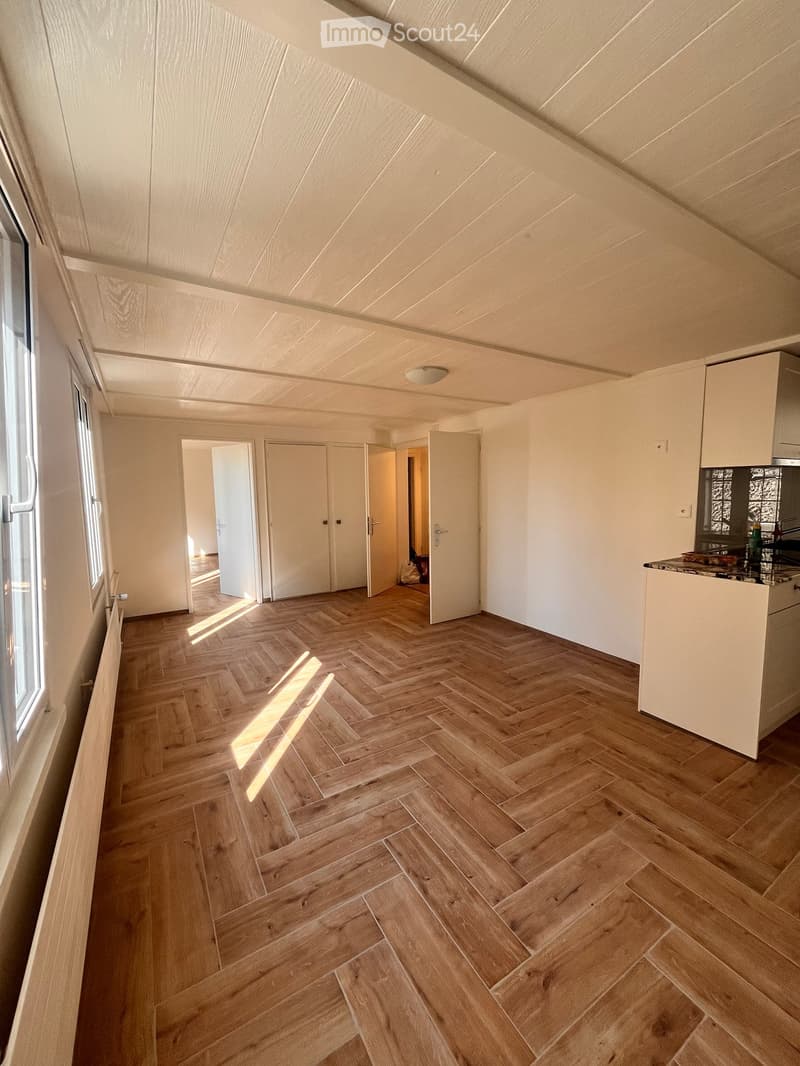 An empty room with wooden herringbone flooring, white walls, and a slanted ceiling. The room features a large window on one side and several doors leading to other spaces.