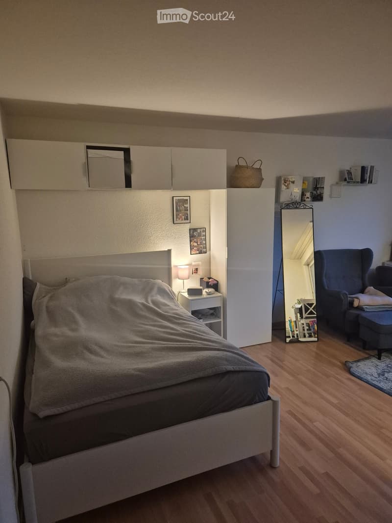 Cozy bedroom with a neatly made bed, nightstand with lamp, mirror, and a blue armchair with ottoman. Wooden flooring and built-in storage units.