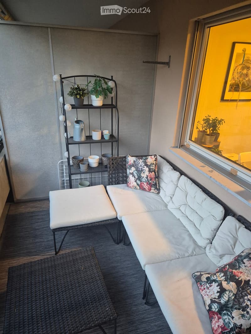 A cozy balcony with a white sofa, floral cushions, a wicker chair, a small table, and a shelf with plants and cups.