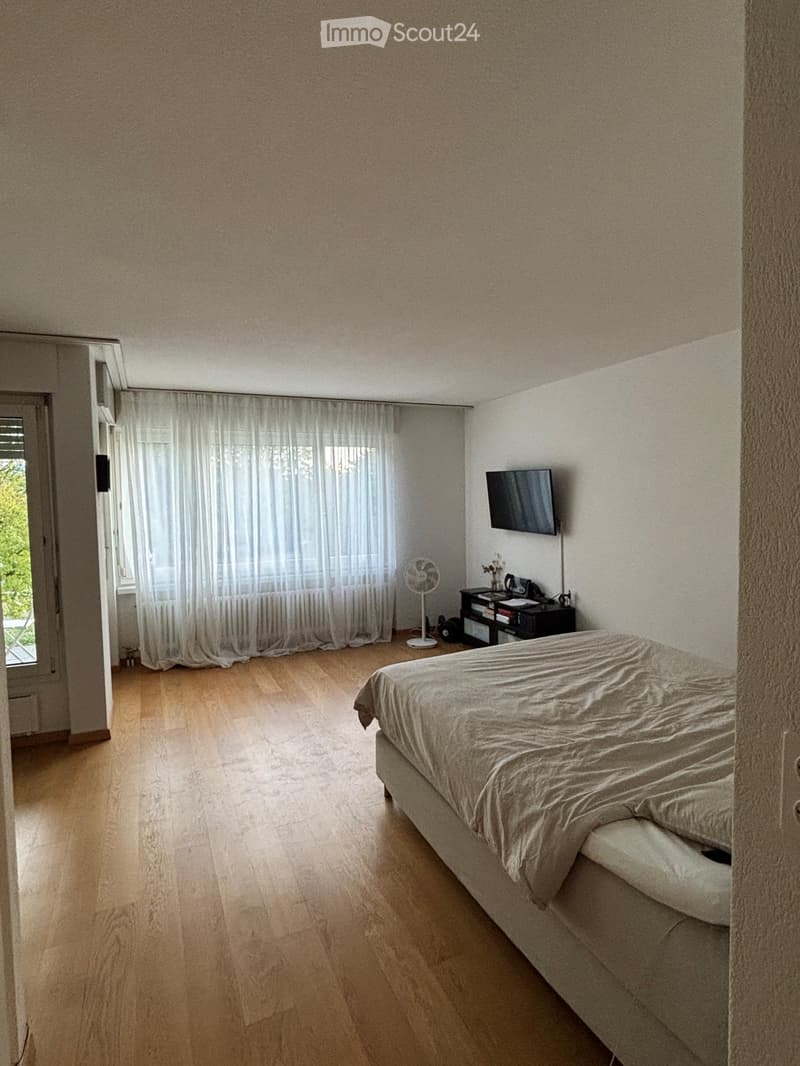 A well-lit bedroom with wooden flooring, a neatly made bed, a nightstand with a fan, a television mounted on the wall, and sheer curtains covering the window.