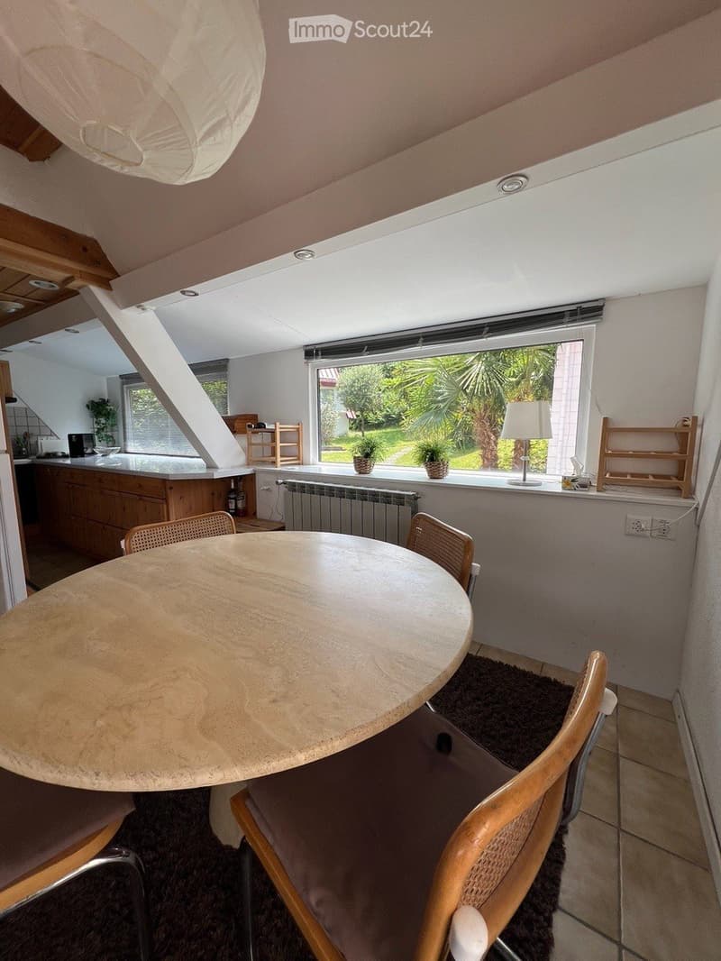 Bright dining area with a round wooden table, four chairs, a lamp on the windowsill, and a view of a garden through large windows. The space includes a kitchen area with cabinets and a counter.