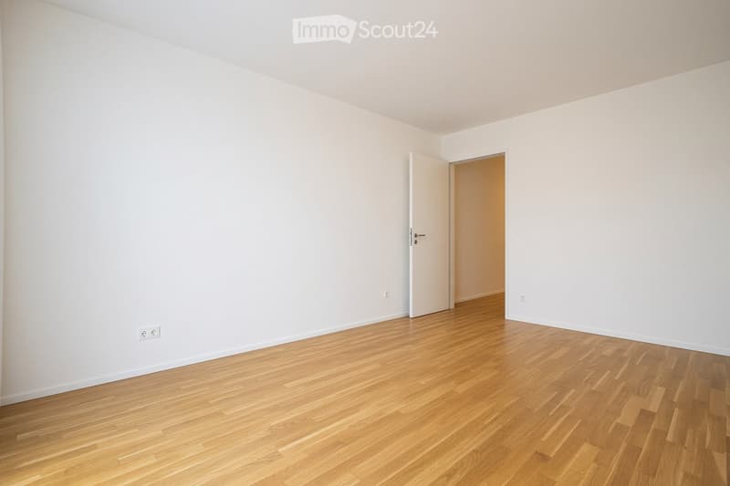 Empty room with light wooden flooring and white walls, featuring an open doorway leading to another room.