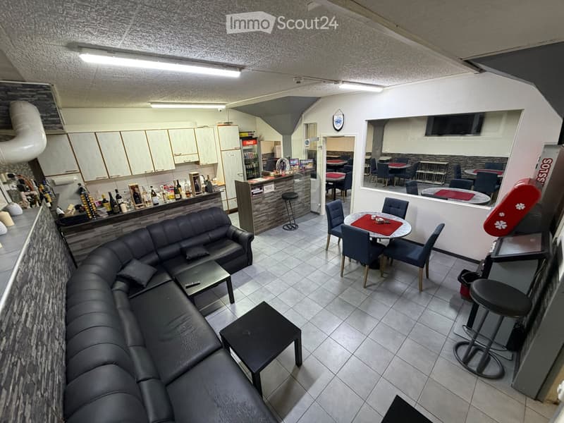 Modern furnished room with a black leather sofa, coffee table, bar area with stools, dining table with chairs, and a refrigerator in the background.
