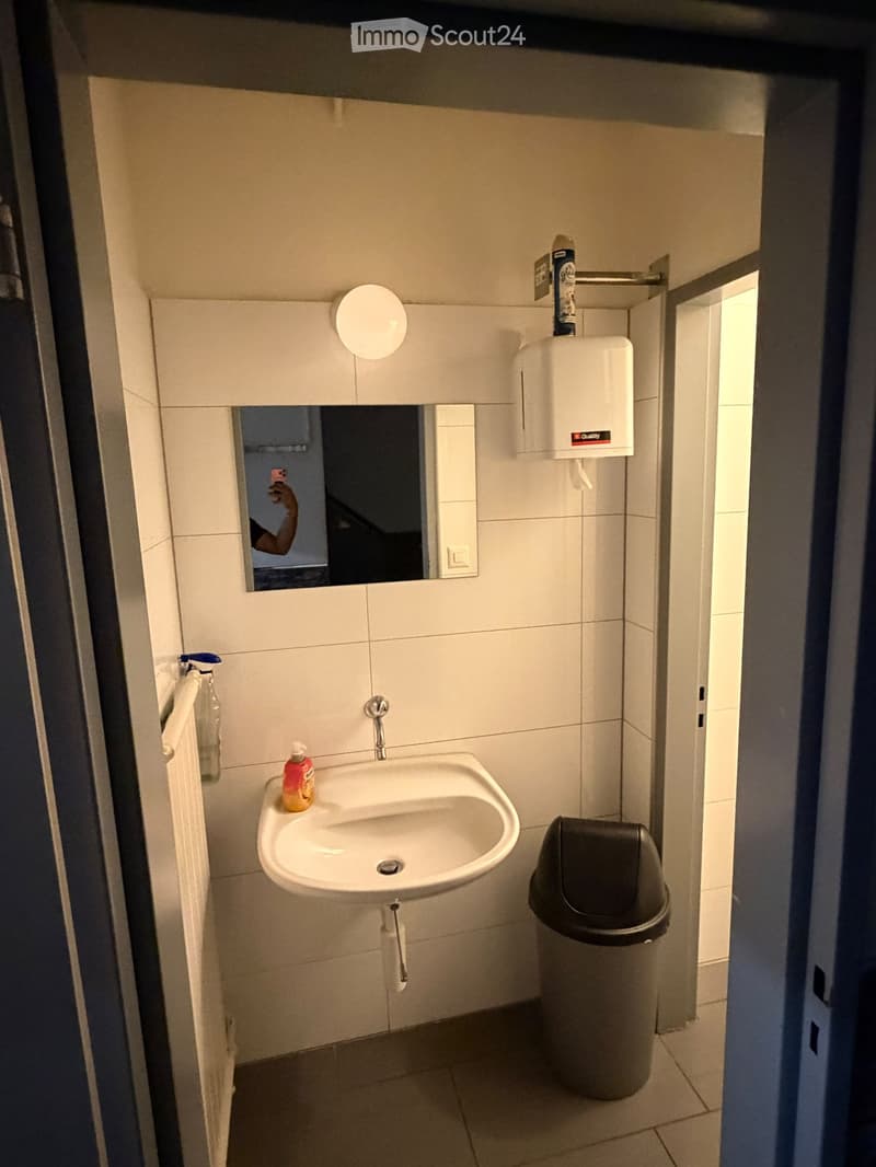 Small bathroom with a white sink, mirror, hand dryer, soap dispenser, and a trash can. The walls are tiled in white, and the floor is tiled in a light color.