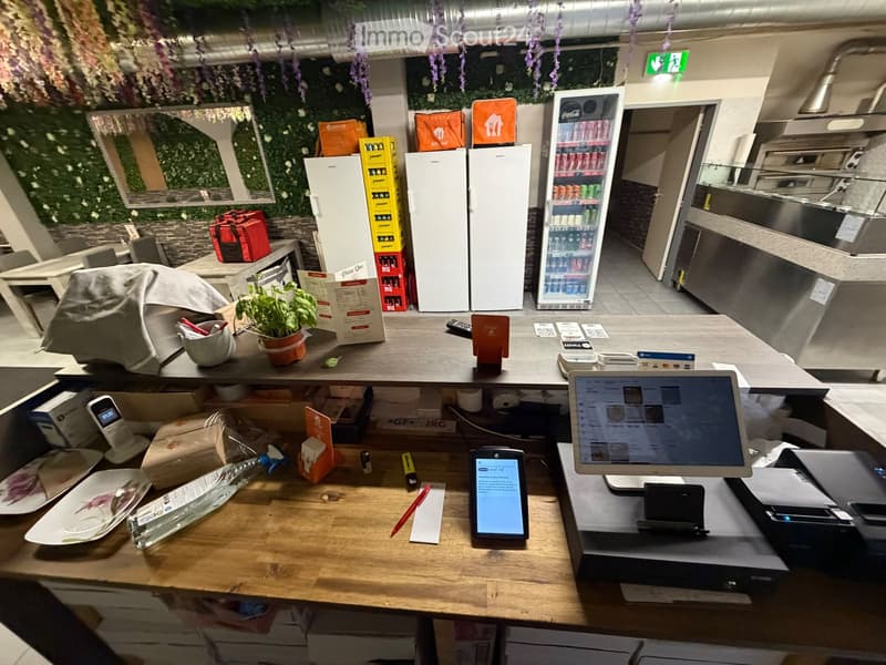 A modern restaurant counter area with a laptop, cellphone, cash register, and several refrigerators in the background. The counter has a potted plant, menu, and various items including a cellphone and a laptop.