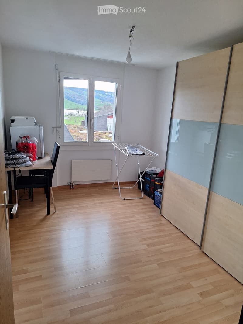 A furnished room with wooden flooring, a large window with a scenic view, a chair, a table with a washing machine, and an ironing board.
