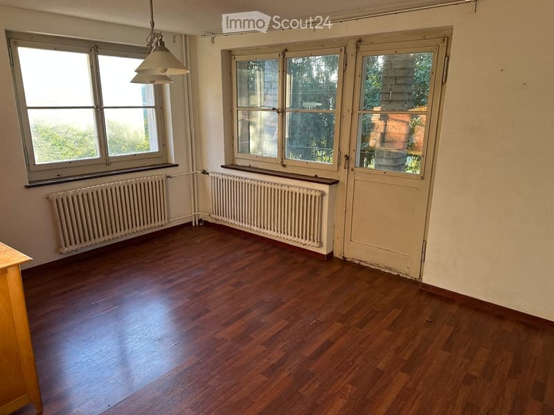Empty room with wooden flooring, large windows, radiators, and a hanging light fixture.