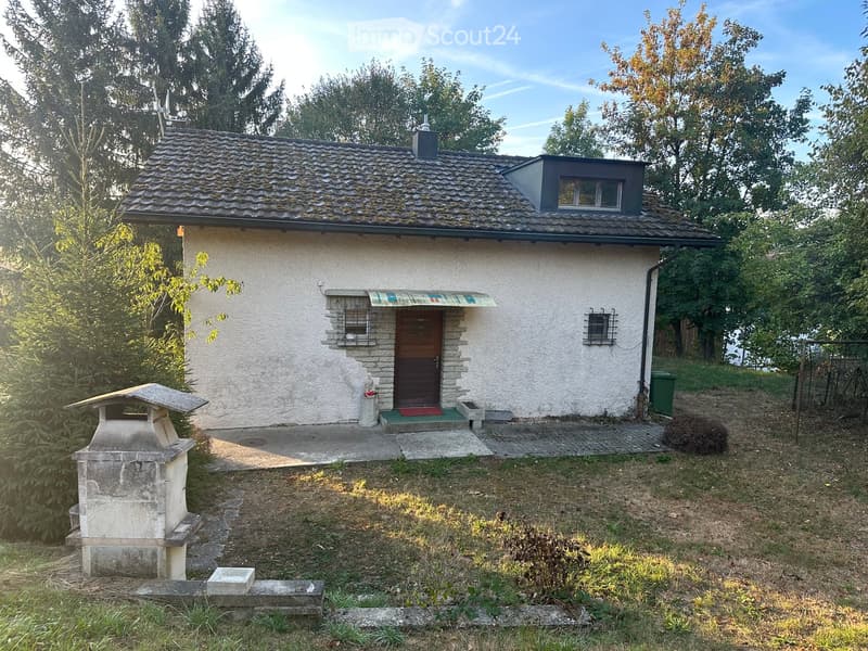 Single-story house with white exterior, dark roof, small dormer window, and a covered entrance with a colorful awning. The yard includes a stone bench, a small statue, and a stone structure resembling a well.