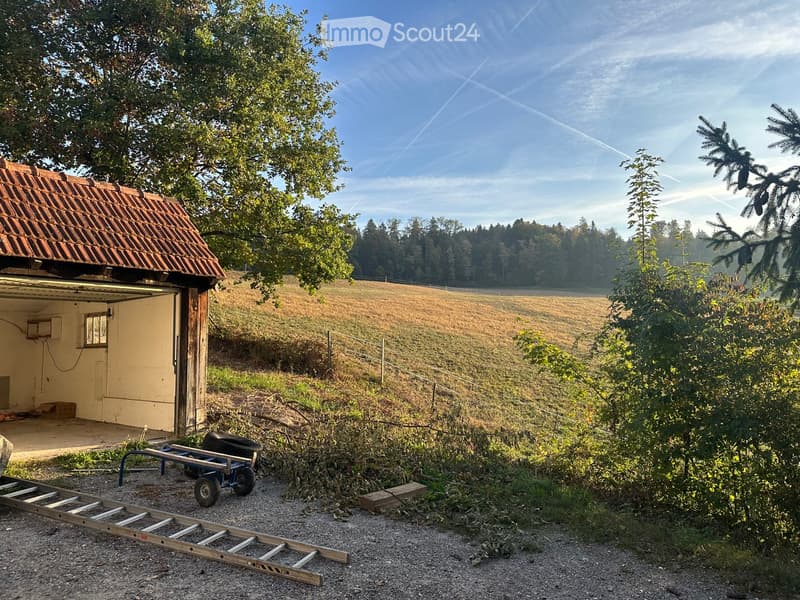 A small, rustic building with a tiled roof and open garage door, situated in a rural area with a field and trees in the background. A ladder and a cart are placed on the gravel ground in front of the building.