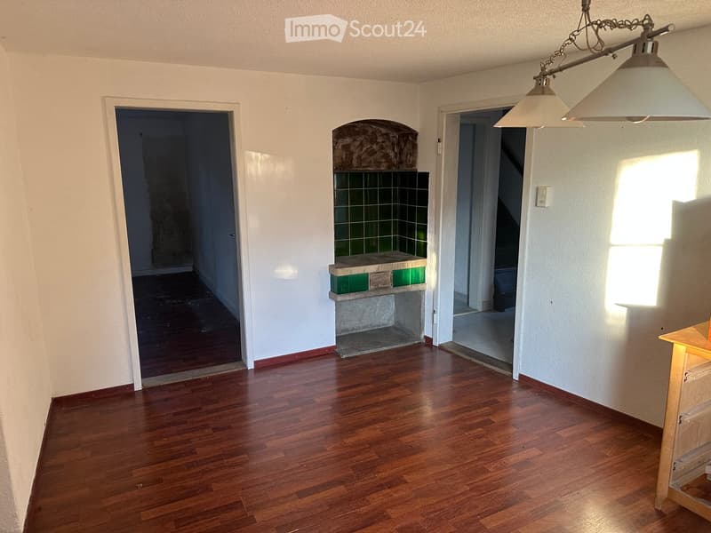 Empty room with wooden flooring, white walls, and a fireplace in the corner. Natural light streams in from a window on the right side.