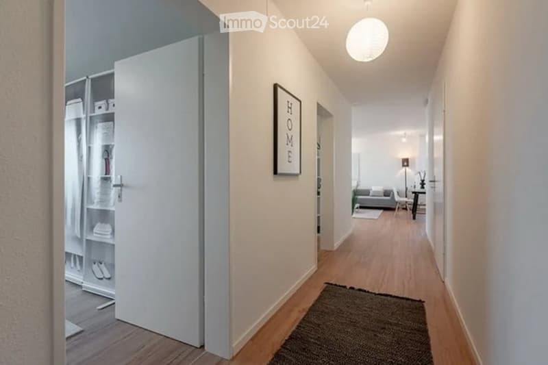 Modern hallway with light wooden flooring, white walls, a round ceiling light, and a framed "HOME" sign. An open closet with shelves and a small rug are visible.