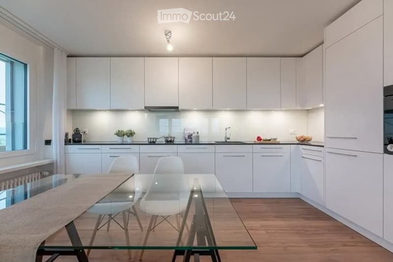 Modern kitchen with white cabinets, a glass dining table, and white chairs. The kitchen features a sink, faucet, and a refrigerator, with a small plant on the counter.