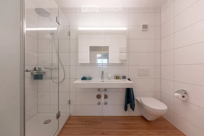 Modern bathroom with white tiles, a glass-enclosed shower, a sink with a mirror, a toilet, and a towel hanging on the wall.