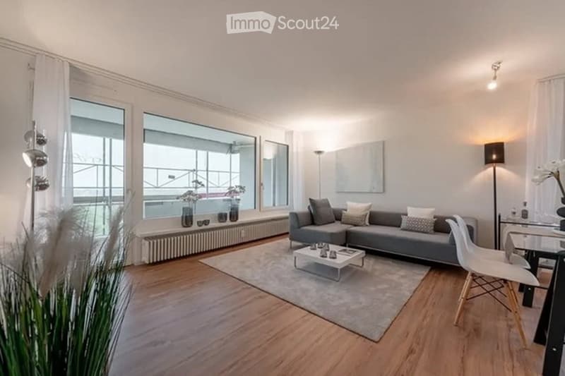 Modern living room with large windows offering a view, grey sectional sofa, coffee table, dining chair, decorative vases, and a floor lamp.