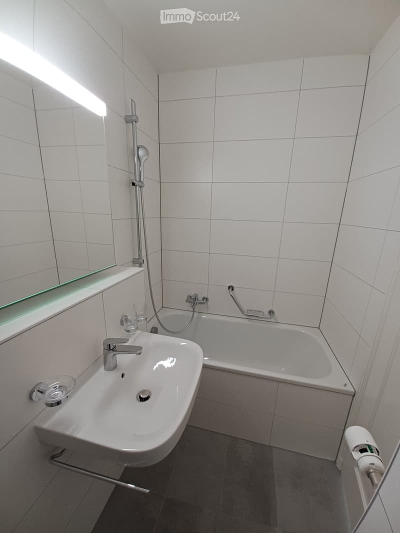 Modern bathroom with white tiles, a sink with a modern faucet, a bathtub with a handheld shower, a mirror, and a cup on the sink counter.