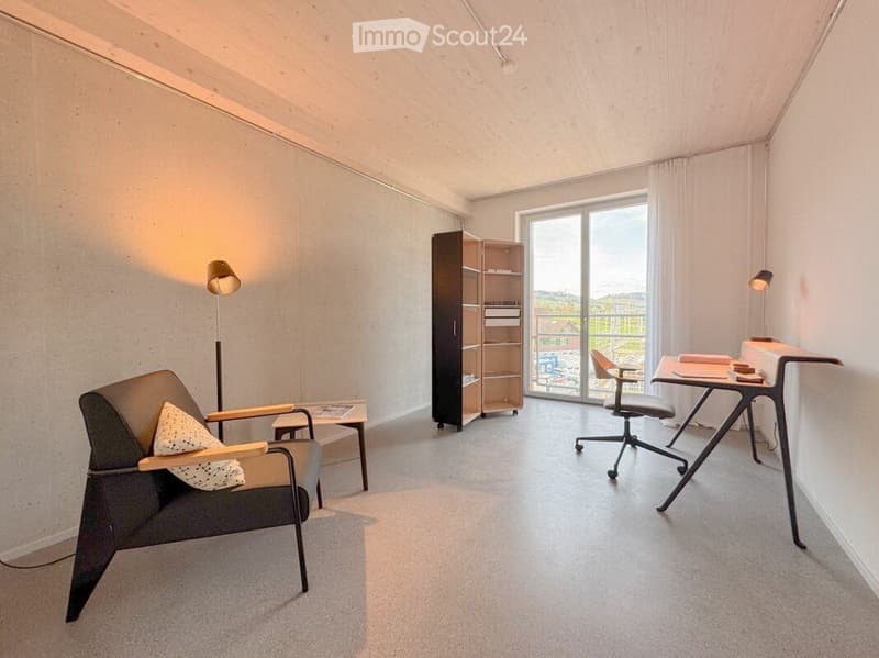 Modern, minimalist room with a grey armchair, small table, shelving unit, desk, and a view through large windows to a balcony.