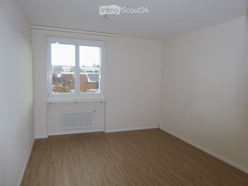 Empty room with wooden flooring, white walls, and a large window with a view of the outside. A radiator is mounted on the wall below the window.
