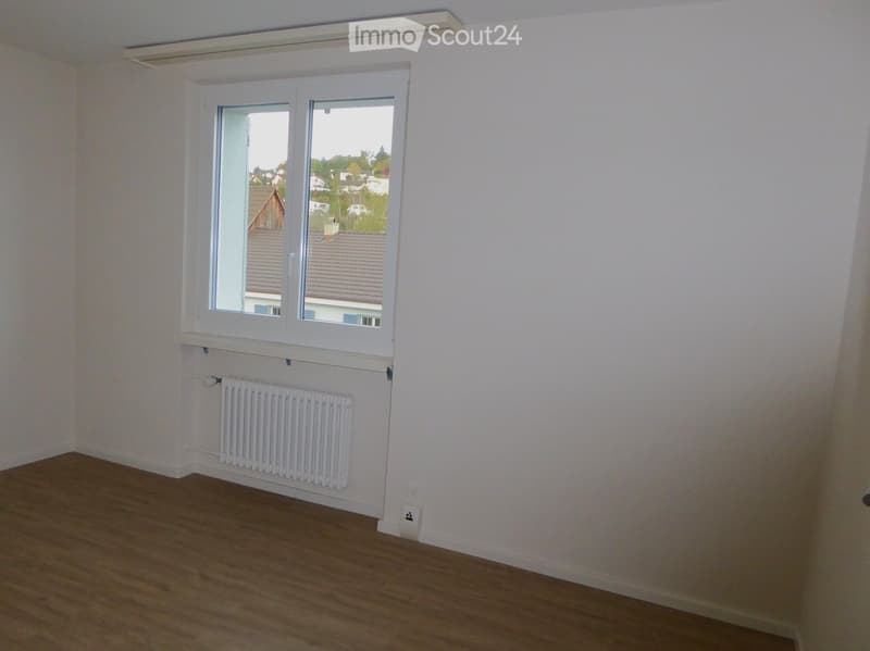 Empty room with wooden flooring, white walls, a window with a view of rooftops, and a white radiator below the window.