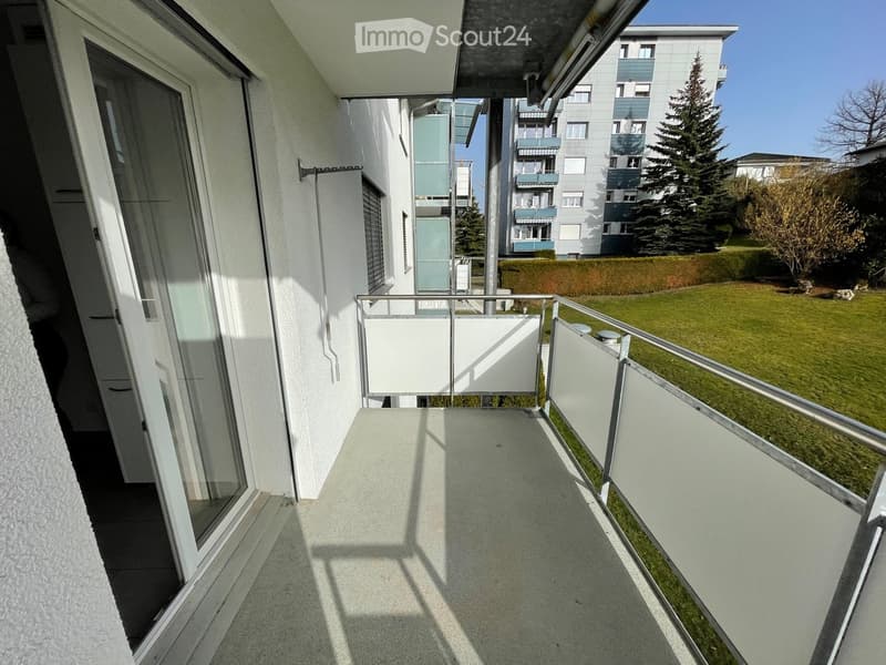 Balcony with modern glass railing, tiled floor, and view of a green lawn and neighboring buildings.