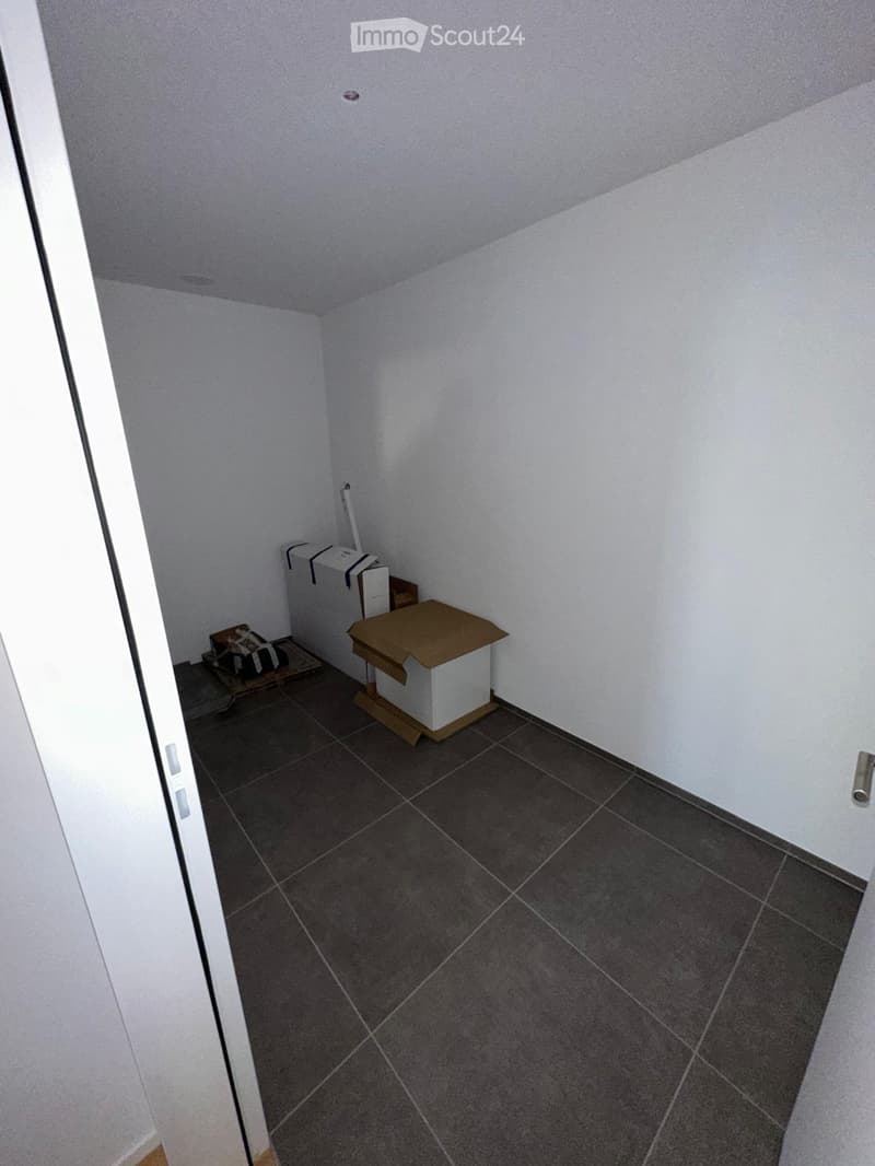 Empty room with white walls, dark tiled floor, and a partially open door on the left side. There are no visible furnishings or decorations.