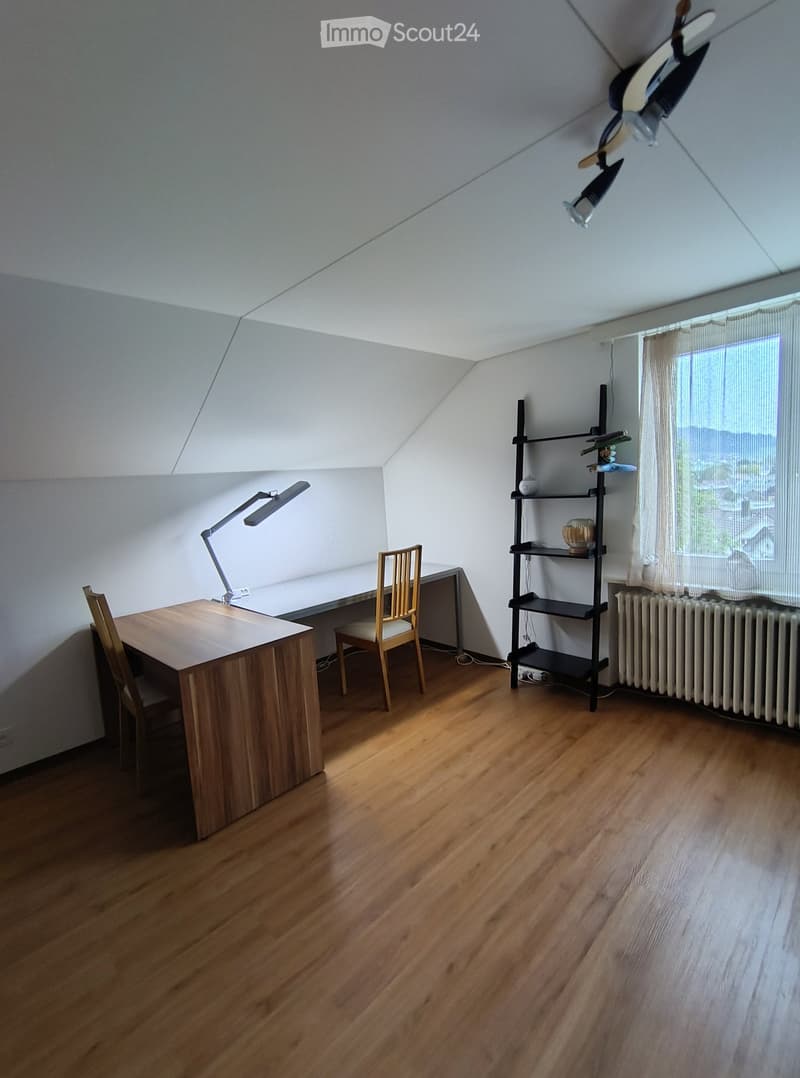 Bright, wooden-floored room with a slanted ceiling, featuring a wooden desk, chair, desk lamp, and a black shelving unit with decorative items.