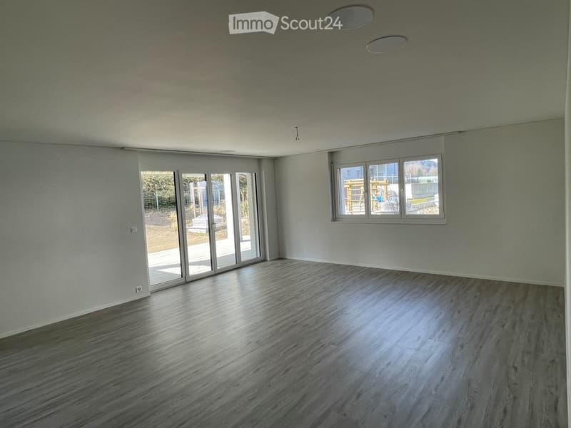 Empty room with light grey walls, polished wooden flooring, large sliding glass doors, and a window with a view of the outdoor area.