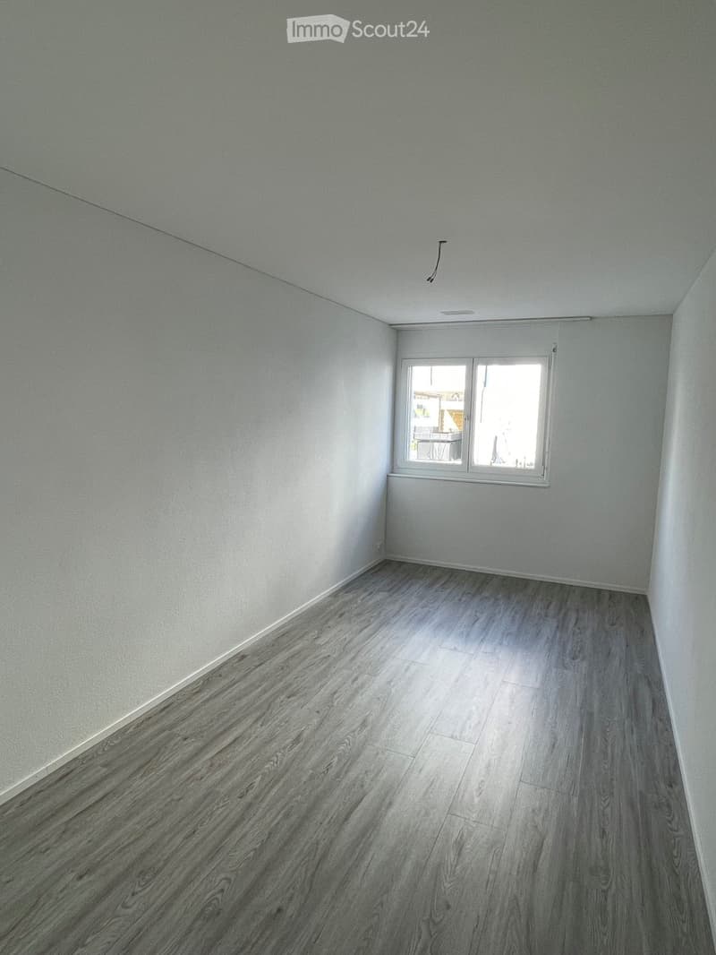 Empty room with light grey walls and wooden flooring, featuring a single window with white frame and blinds.