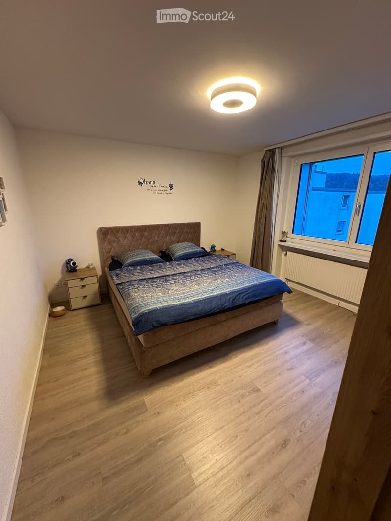 Cozy bedroom with wooden flooring, large window with curtains, bed with blue bedding, nightstand with lamp, and decorative items.