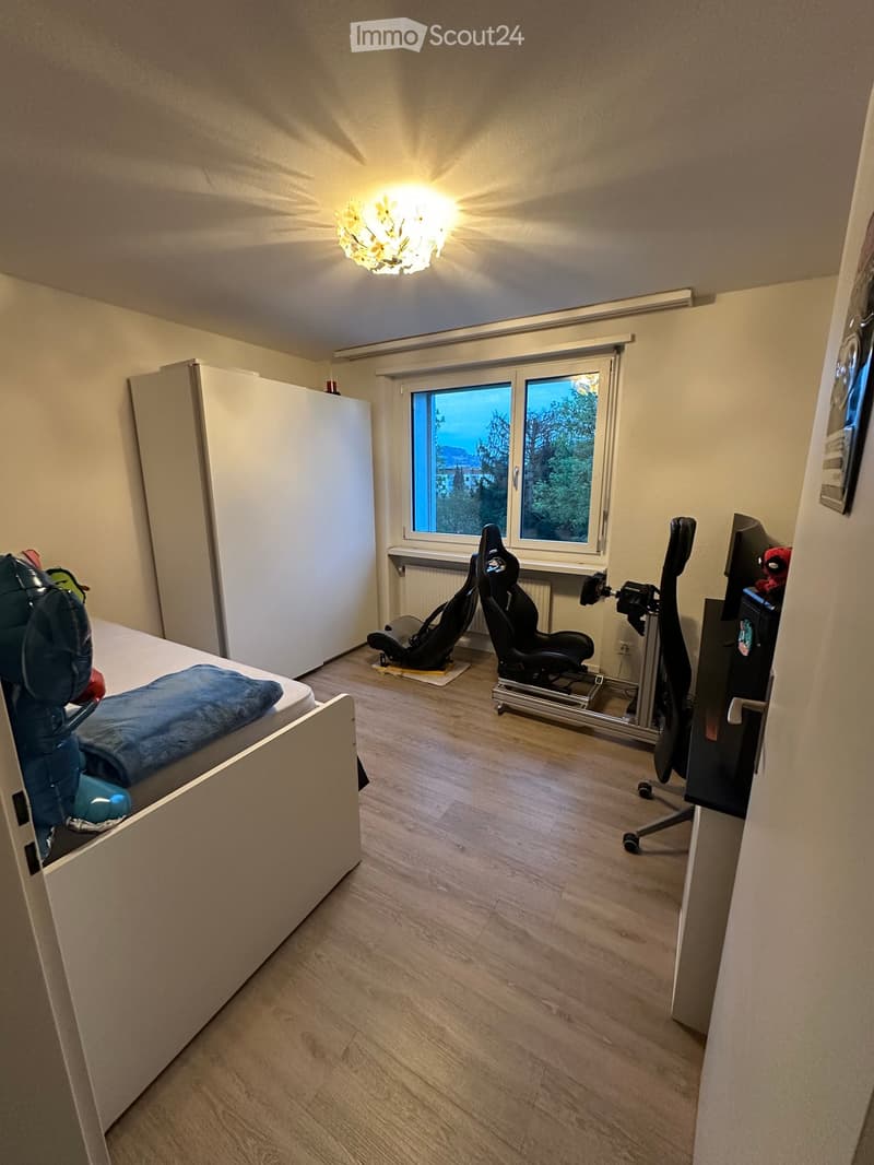 A furnished room with a bed, two chairs, a screen, a table, and a cupboard. The room has wooden flooring and a window with a view of trees.