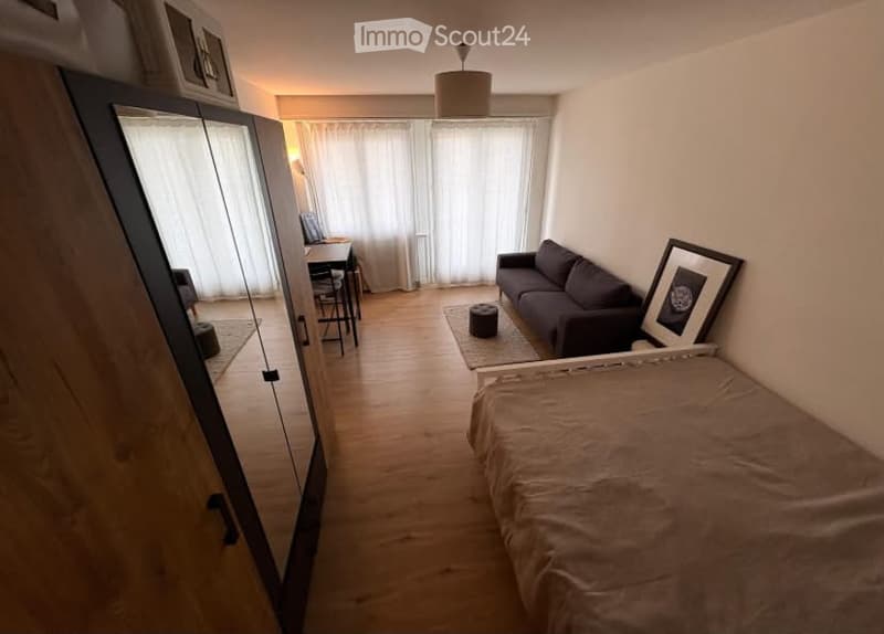 A compact, well-lit room with a bed, sofa, wardrobe, and a small table with chairs. The space features wooden flooring and large windows with white curtains.