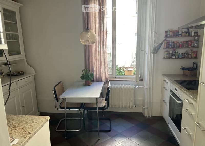 A compact kitchen with a small dining area. Features include a white table with two chairs, a potted plant, built-in cabinets, an oven, and shelves with various items.