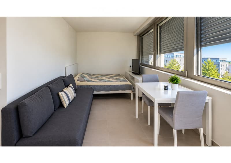 A modern, compact living space with a sofa, bed, small table, chairs, and a TV screen, featuring large windows with blinds and a view of the city.