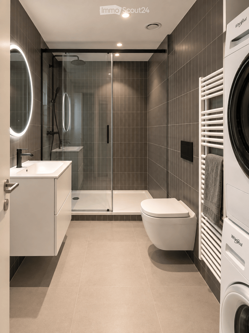 Modern bathroom with dark brown tiles, a white sink with a sleek faucet, a large mirror with LED lighting, a glass-enclosed shower, a toilet, a radiator with a towel, and a washing machine.
