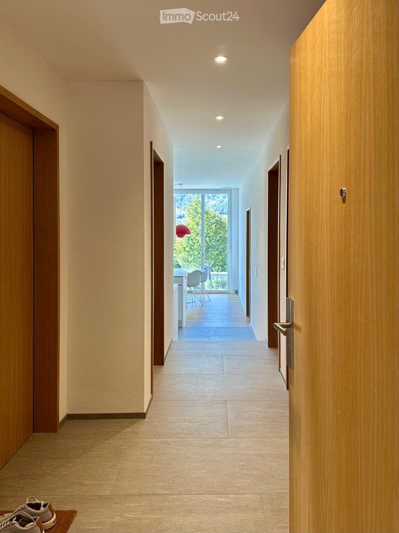 Modern hallway with light-colored tiles, wooden paneling, recessed ceiling lights, and a view of a dining area through a large glass door.