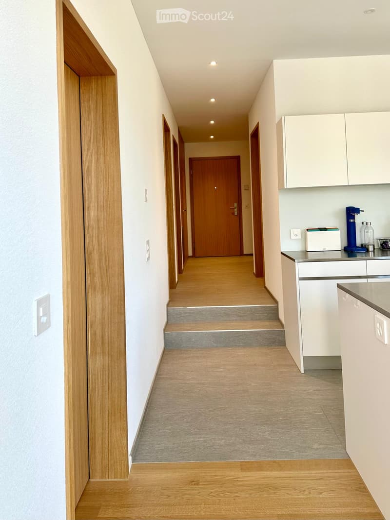A modern hallway with wooden flooring, white walls, and recessed lighting. A small kitchen area is visible on the right with white cabinets, a countertop, and a few appliances.