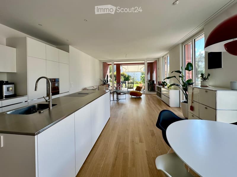 Modern kitchen with white cabinets, stainless steel sink, and a dining area with a round table and chairs. The space features wooden flooring and large windows leading to a patio.