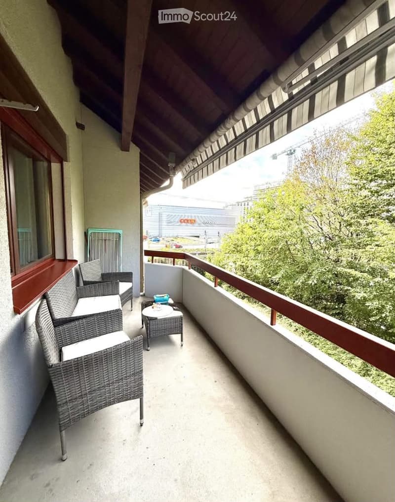 Balcony with wicker chairs, small table, and a view of greenery and a building.