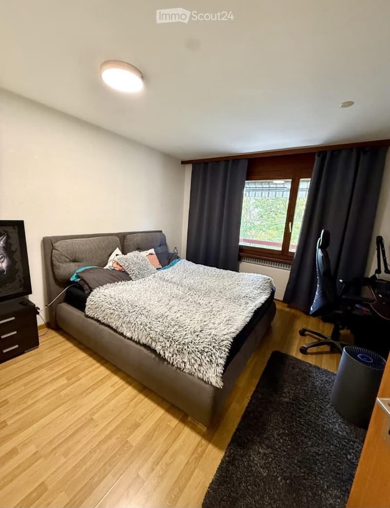 A modern bedroom with a large bed, a desk, a chair, and a TV screen. The room features wooden flooring, large windows with dark curtains, and a cozy, well-lit atmosphere.