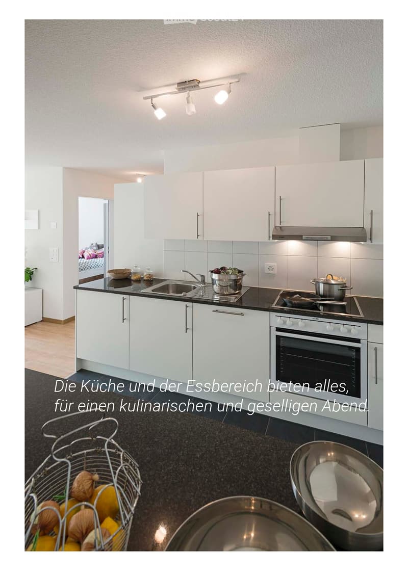 Modern kitchen with white cabinets, black countertops, stainless steel appliances, and a sink. Visible items include a pot, pan, bowl, and a basket with fruits.