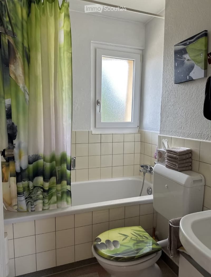 Bathroom with a bathtub, toilet, sink, and a green bamboo-patterned toilet seat cover. The room features tiled walls and a window with a curtain.
