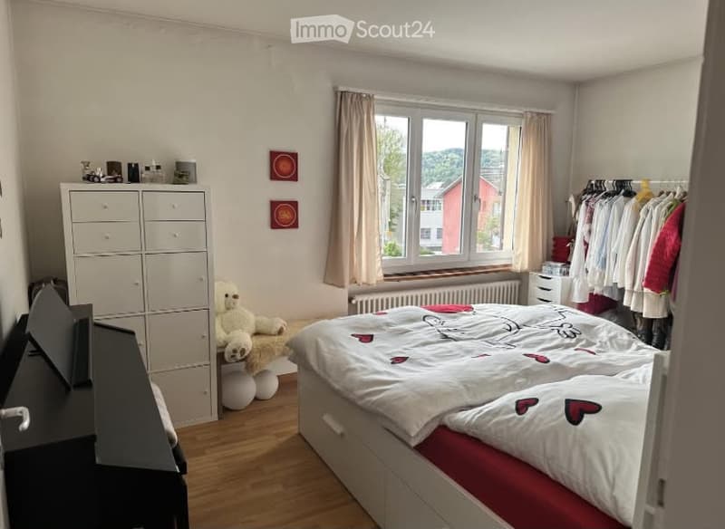 Cozy bedroom with a double bed, white wardrobe, a teddy bear on a stool, and a clothing rack with hanging clothes. The room features wooden flooring and large windows with curtains, offering a view of the outside.