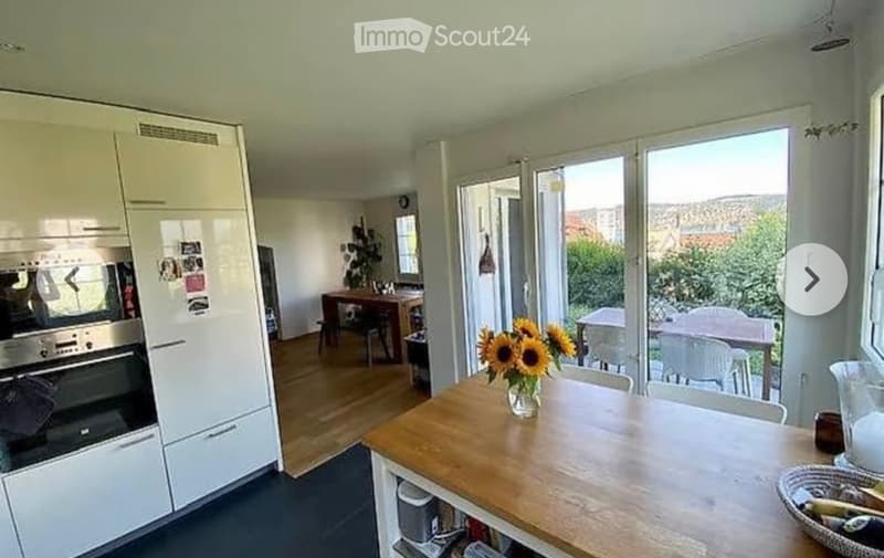 A modern, well-lit kitchen and dining area with white cabinets, a wooden dining table, and a vase of sunflowers on the table. Large windows provide natural light and a view of the garden.