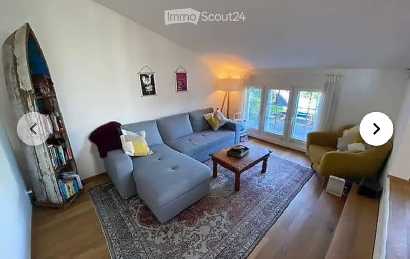 Bright living room with a grey sectional sofa, wooden coffee table, armchair, floor lamp, and a unique wooden bookshelf. Large windows provide natural light.