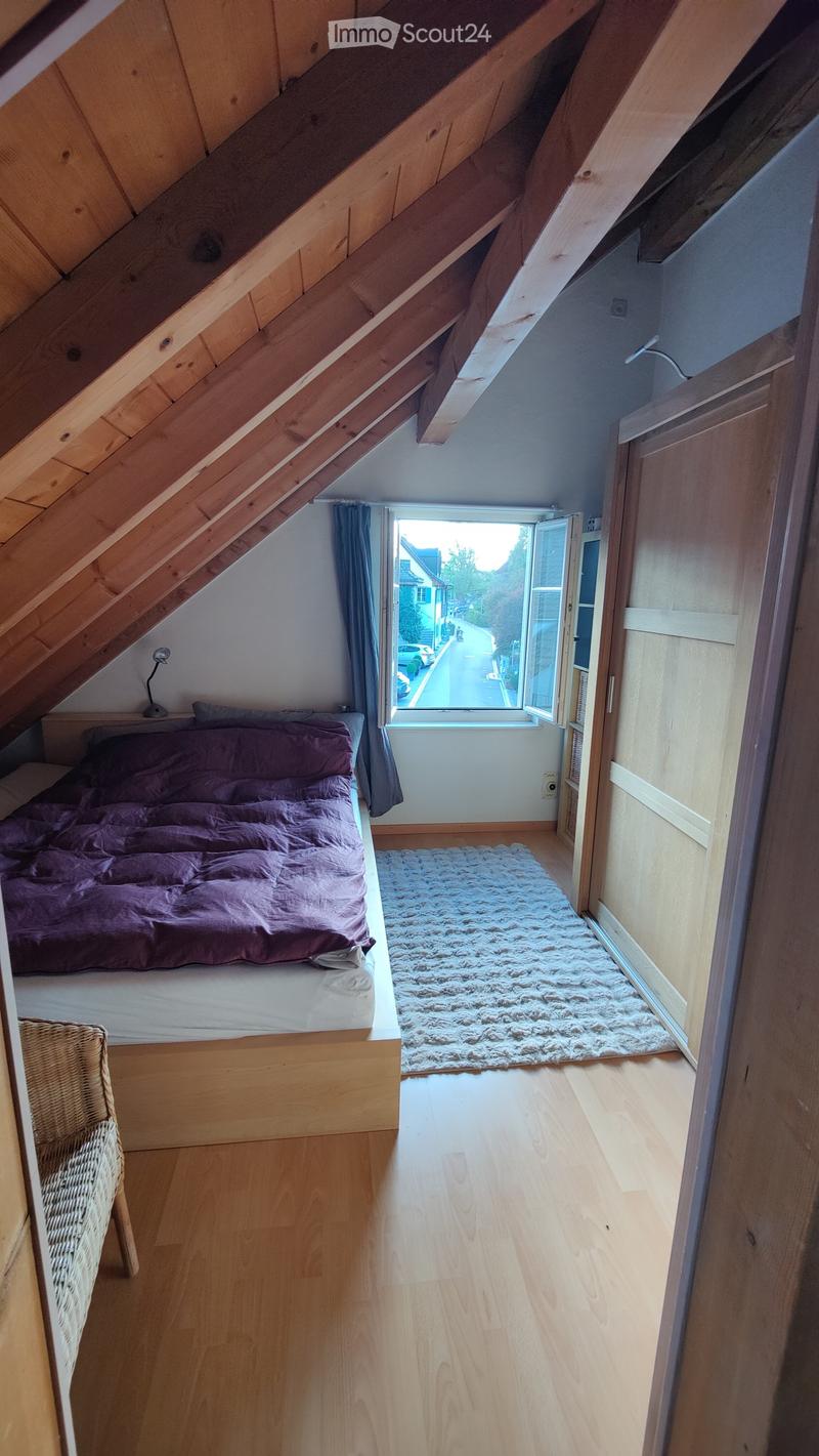 Cozy attic bedroom with wooden slanted ceilings, a single bed with purple bedding, a wicker chair, a fluffy white rug, and a window with blue curtains overlooking a street.