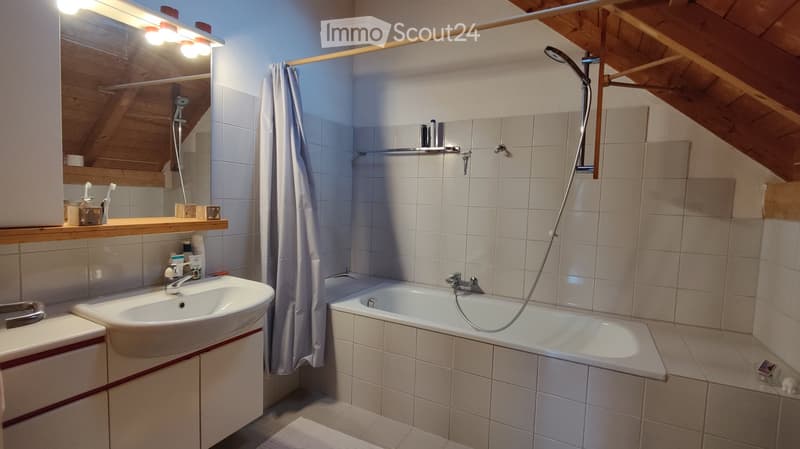 Cozy bathroom with a sink, bathtub, and shower area. Features a wooden shelf with toiletries, a mirror with built-in lighting, and a slanted wooden ceiling.