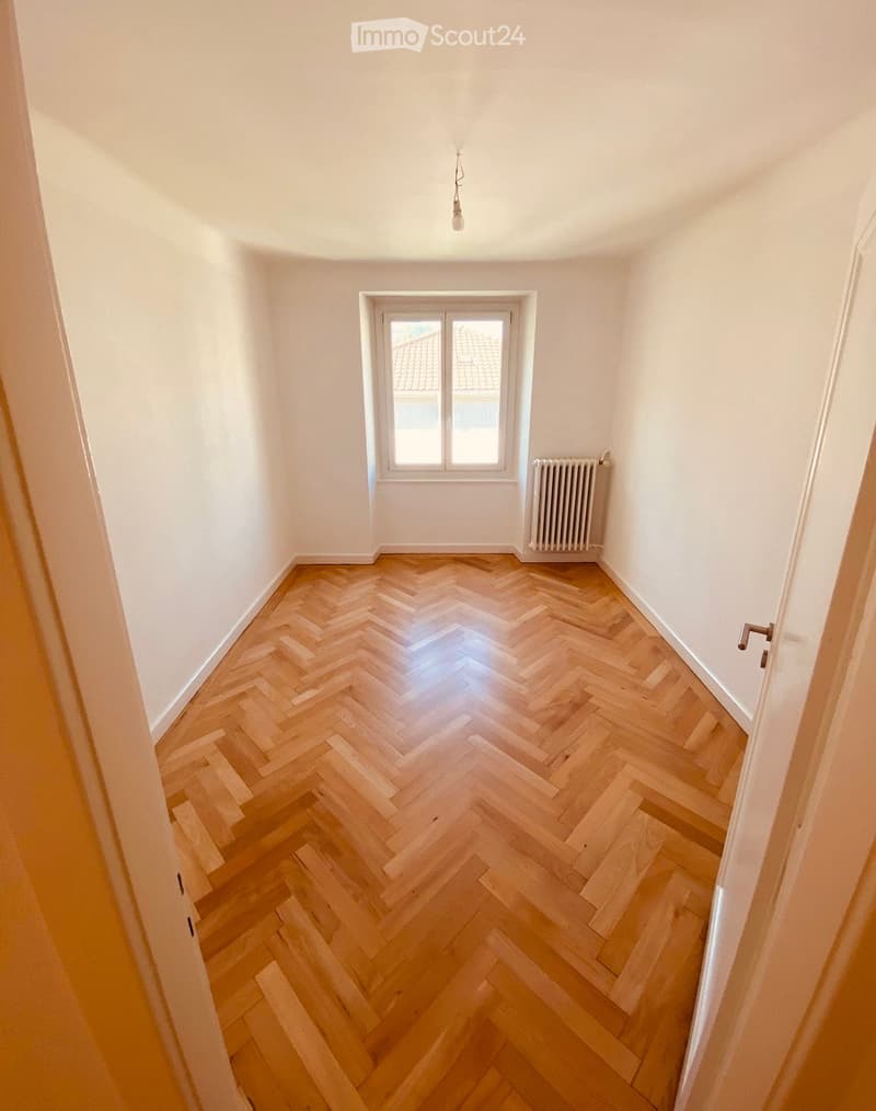 Empty room with herringbone wooden flooring, white walls, a single window, and a radiator. No furniture or personal items are present.