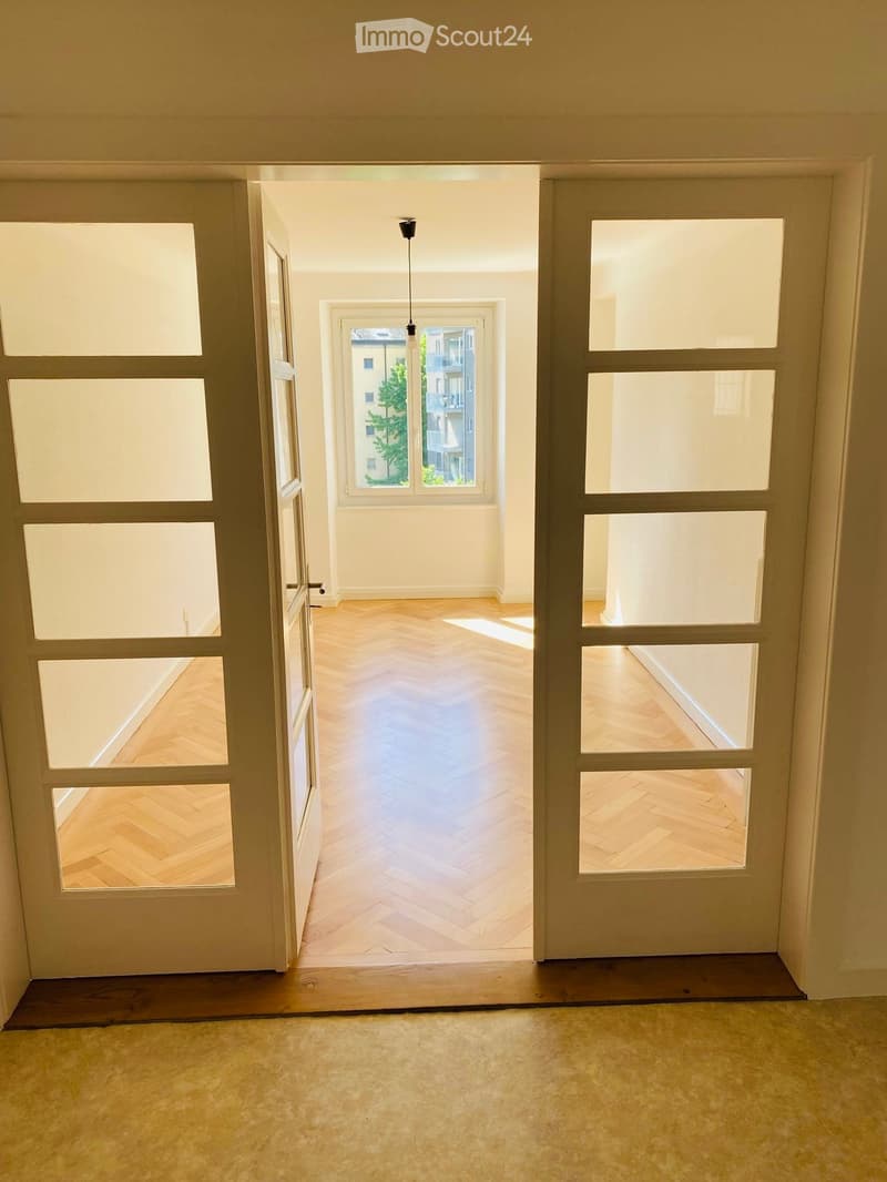 Empty hallway with wooden flooring and large windows, leading to another room with wooden herringbone flooring and natural light.