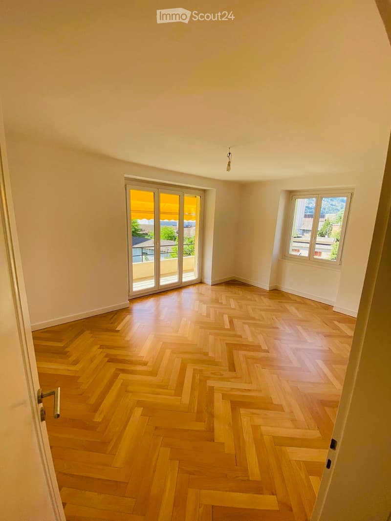 Empty room with light wooden herringbone flooring, large sliding glass doors leading to a balcony, and a window with white frames.