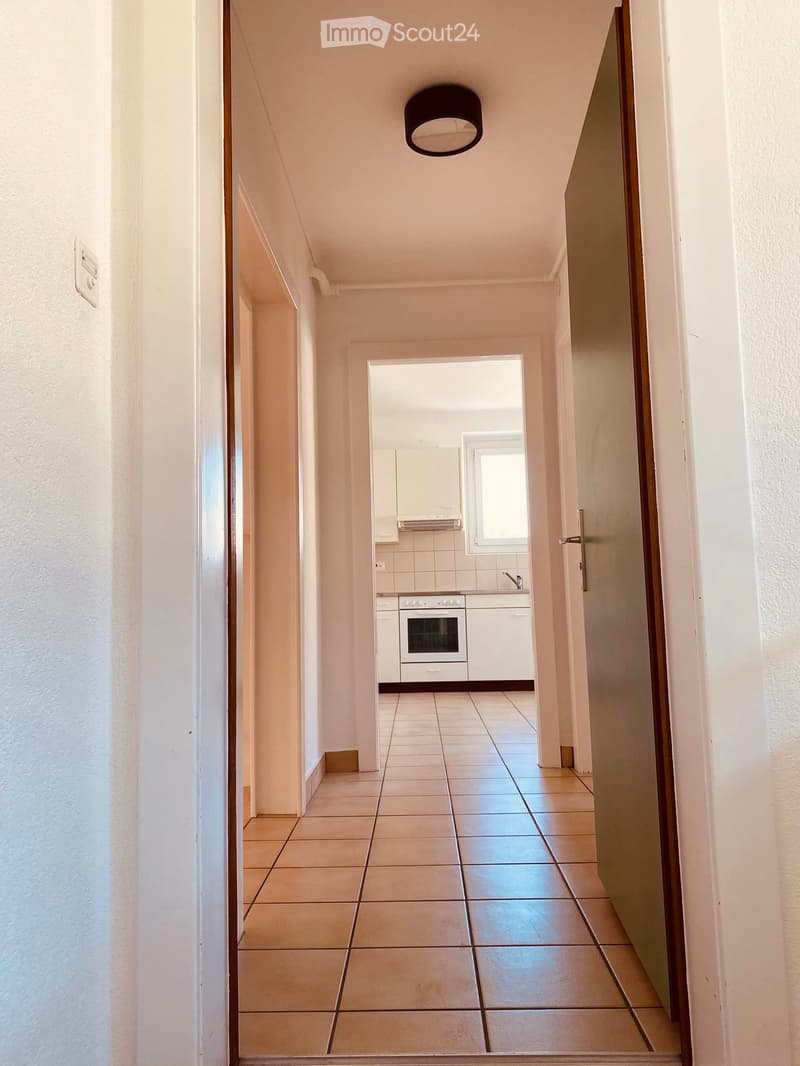 A narrow hallway with tiled flooring, white walls, and a ceiling light fixture. A door on the right leads to a kitchen area with white cabinets, a built-in oven, and a window.